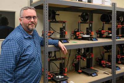 Man in a blue plaid shirt leans on metal shelves holding multiple Prusa 3D printers.