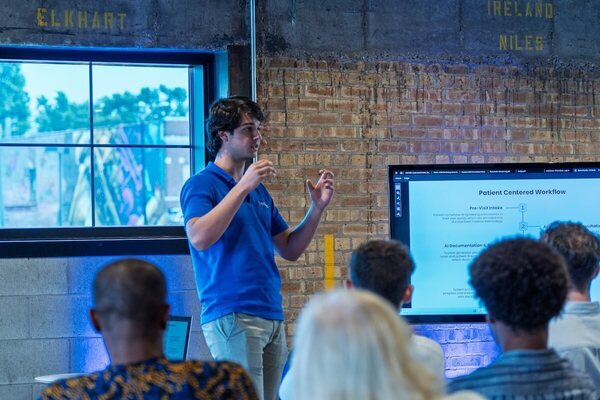 A presenter in a blue shirt gestures while speaking to a small audience in front of a screen displaying "Patient Centered Workflow".