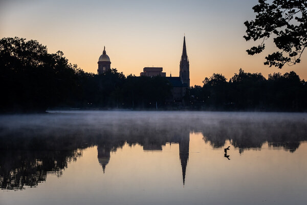Golden Dome and Basilica of the Sacred Heart silhouetted against an orange-blue sky, reflected in misty St. Mary's Lake at twilight. Dark trees line the bank, with a bird floating on the water.