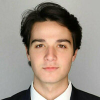 Young man with dark hair and brown eyes wearing a dark suit jacket and white shirt, looking forward directly.