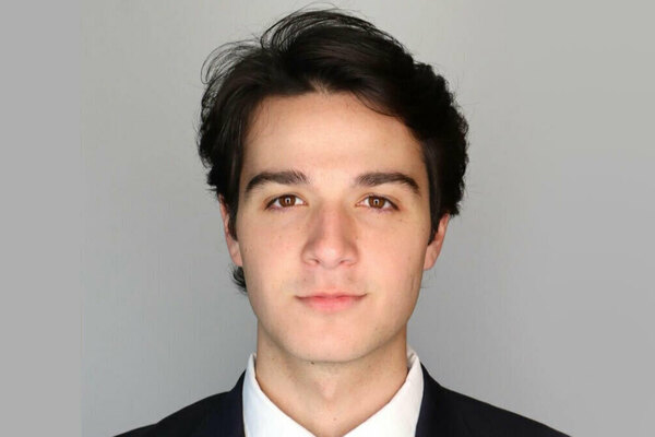 Young man with dark hair and brown eyes wearing a dark suit jacket and white shirt, looking forward directly.
