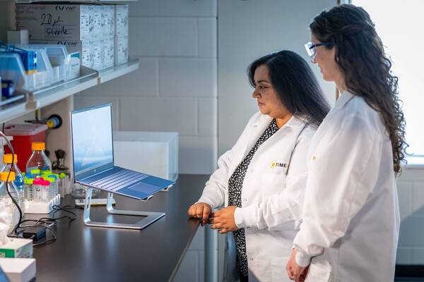 Two researchers in lab coats, one wearing blue glasses, observe an open laptop on a stand in a science lab.