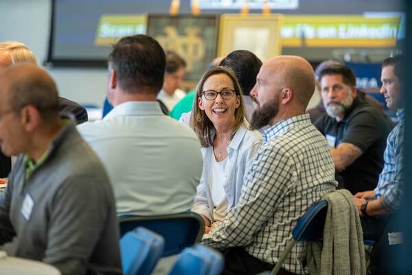Smiling woman in glasses and light blue shirt converses with a bearded man in plaid at a group event.