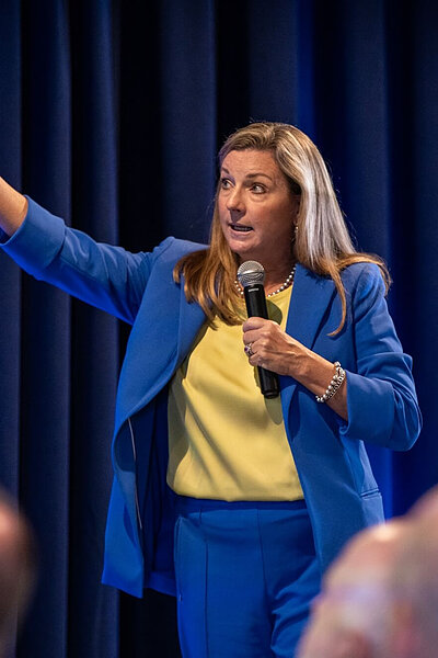 Woman in a blue suit and yellow top speaks into a mic, pointing with a presentation remote against dark blue curtains.