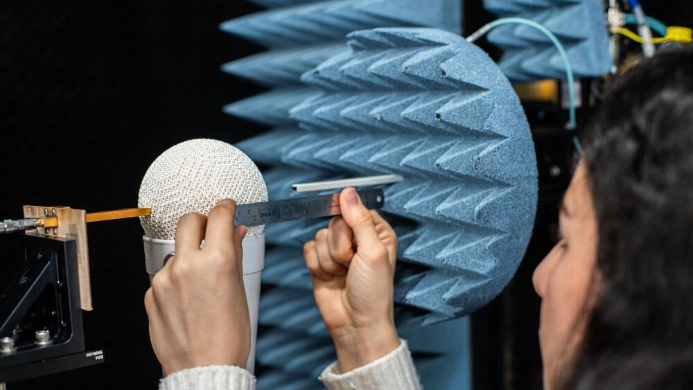 Hands measure a white porous sphere with a metal ruler, positioned against a blue pyramidal anechoic chamber wall.