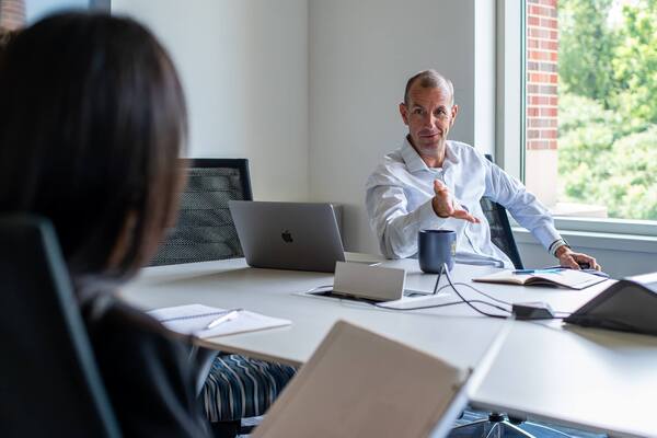 Man in a light blue shirt smiles, gestures at a conference table with a laptop and Notre Dame mug.