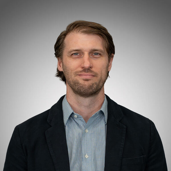 A man with light brown hair, a beard, and blue eyes smiles slightly, wearing a dark blazer over a blue checkered shirt.