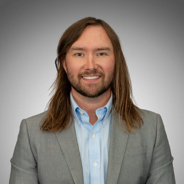 Smiling man with long brown hair and beard wears a light blue gingham shirt and gray suit jacket.