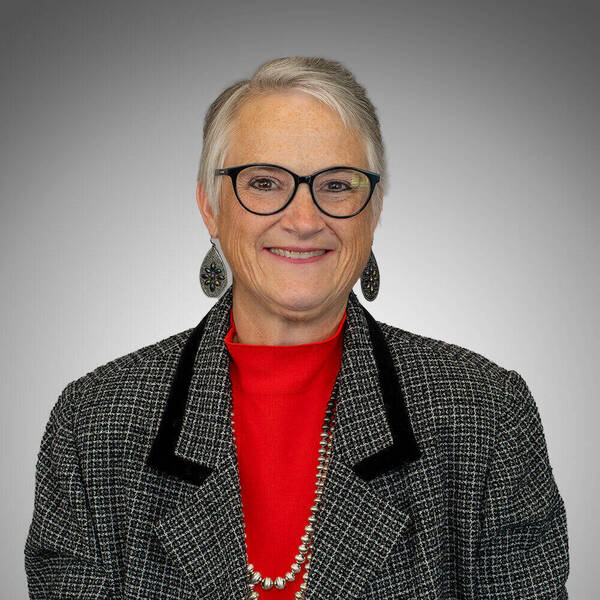 Smiling woman with short grey hair, black framed glasses, a red turtleneck, black and white tweed blazer, and silver beaded necklace.