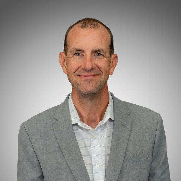 Smiling man with short brown hair, wearing a light gray blazer over a light plaid shirt.