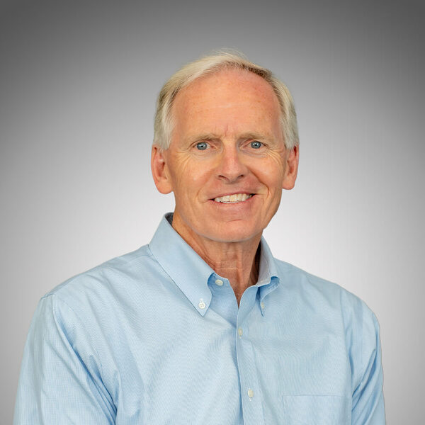 A smiling man with gray hair and blue eyes wears a light blue button-down shirt against a gray gradient background.