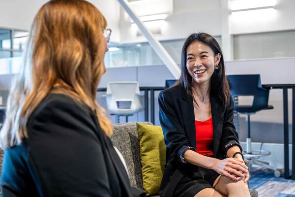 Two women chat in a modern office. One woman, in a black blazer and red top, smiles broadly at the other.