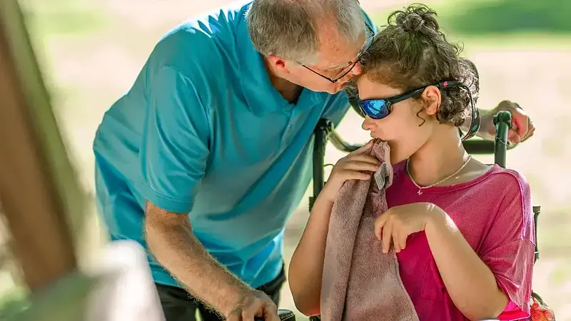 Older man in blue shirt leans toward young person in wheelchair with sunglasses, holding a towel to mouth.