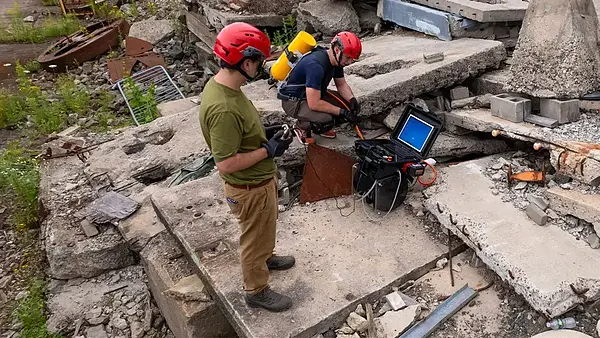 Two people in red helmets operate equipment amongst concrete debris.