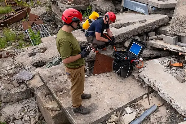 Two people in red helmets operate equipment amongst concrete debris.